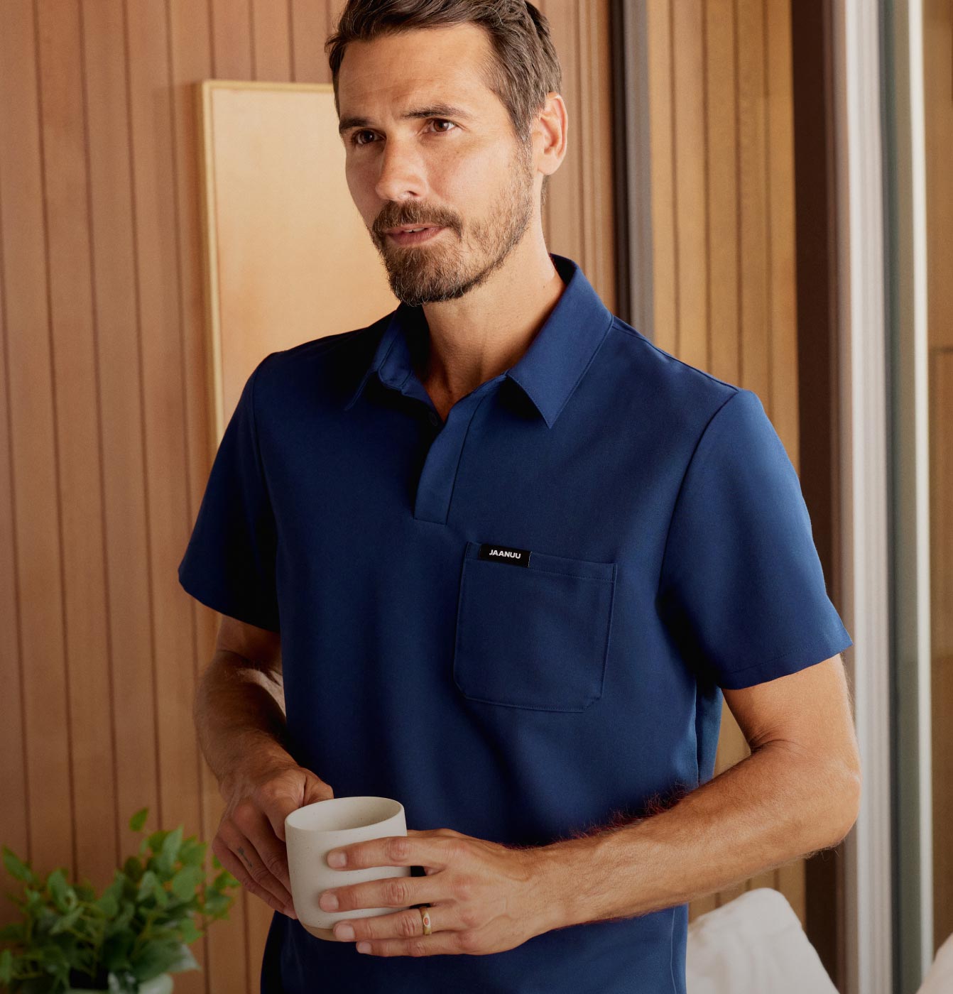 A man wearing a polo scrub top and holding a coffee mug in his hands.