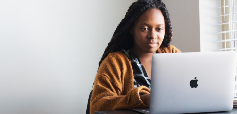 A black woman sits at a table with a laptop. She has long braided hair and wears a yellow sweater.