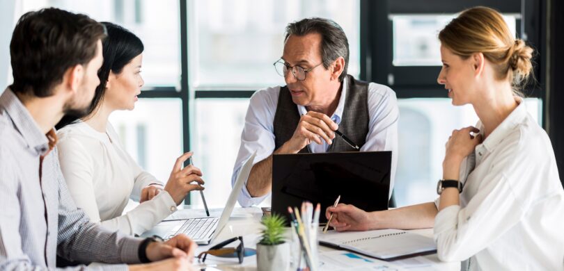 A group of people sit around a table in a brightly lit office. There is one older man, two younger women, and one younger man. They wear professional clothing.