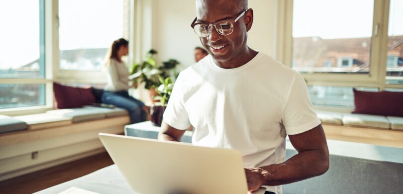 A man sits with a laptop in a brightly lit office setting. He has a shaved head and wears a white tshirt and glasses.