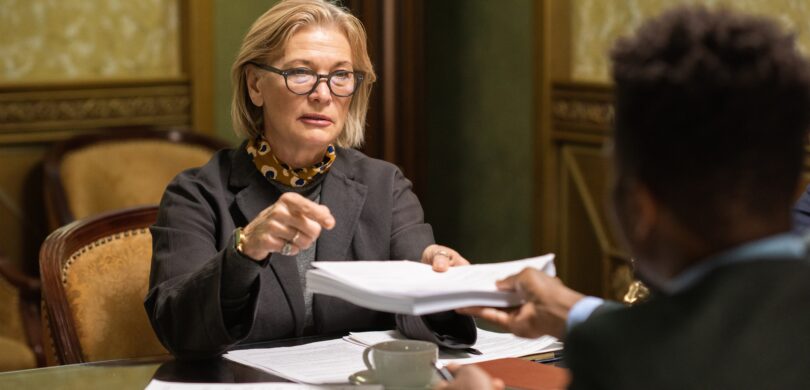 An older woman sits in a business meeting in a luxurious office. She is surrounded by papers.