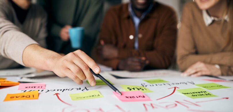 A team of diverse professionals sits in the background. They are looking at a large piece of paper covered in writing and colorful post-it notes.
