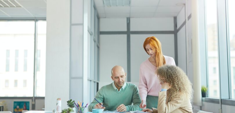 A team of three coworkers sits in a brightly lit office around a table covered in papers and coffee mugs. There is a white man and two women, one white and one of an unidentified race.