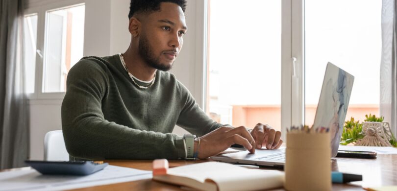 A black man sits at a desk in front of a computer and pads of paper. He wears a dark green long-sleeved shirt and a beaded necklace. He has short, dark hair.