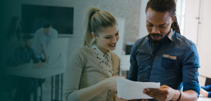 A man and a woman stand together looking at a piece of paper. They are in a busy meeting room with other people in the background. The man is black and the woman is white. They both wear professional clothing.