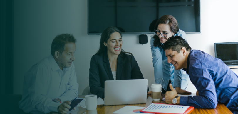 A team of coworkers including two men and two women sit at a table, looking at a laptop.