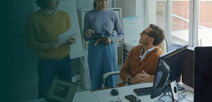 A team of professionals in a busy office setting. There is a teal blue gradient overlay on the image.