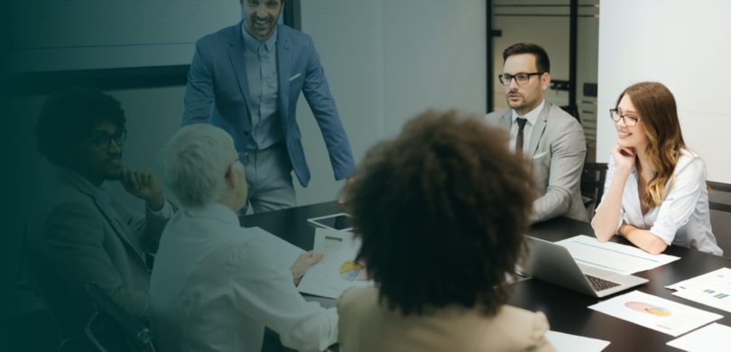 A team of professionals in a meeting. A young man in a blue suit stands at the end of a table in front of a business presentation.